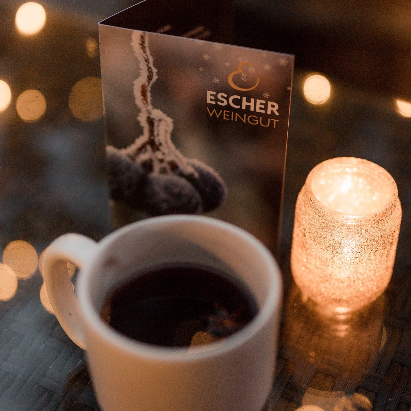 Foto einer weißen Tasse mit ESCHER-Glühwein darin neben einer Kerze und einem Flyer des Weingut Escher Schwaikheim im Hintergrund